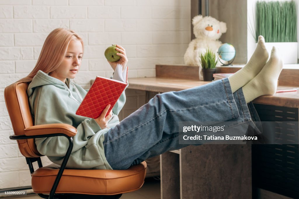 Pretty blonde teenage girl in home clothes reading textbook,doing homework in a relaxed pose at home at her desk.Back to school concept