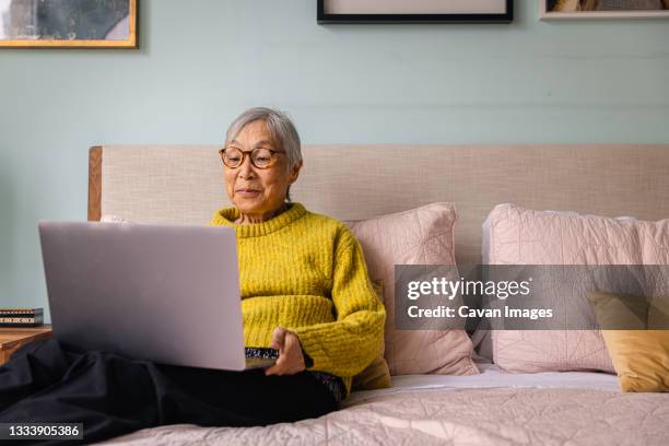 senior asian woman using laptop while sitting in bedroom at home - senior adult on computer stock pictures, royalty-free photos & images