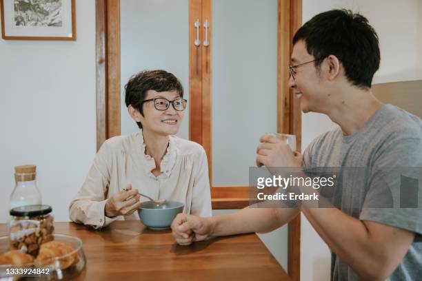 positive asiatische ältere erwachsene frau, die ihrem sohn am esstisch zulächelt, während sie müsli mit milch isst - stockfoto - weekday stock-fotos und bilder