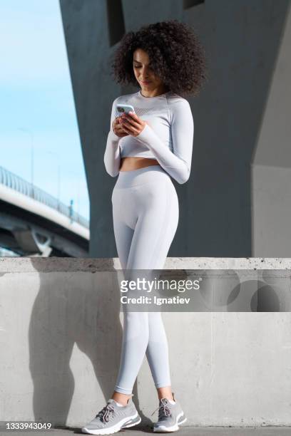 una runner femminile riccia in forma e attraente in un abito da corsa bianco con un supporto a maniche lunghe con messaggi a gambe incrociate al telefono - leggings foto e immagini stock