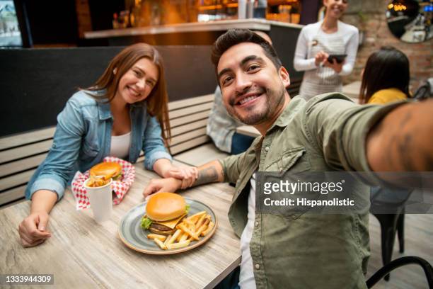 couple heureux prenant un selfie tout en mangeant des hamburgers dans un restaurant - diner amoureux photos et images de collection