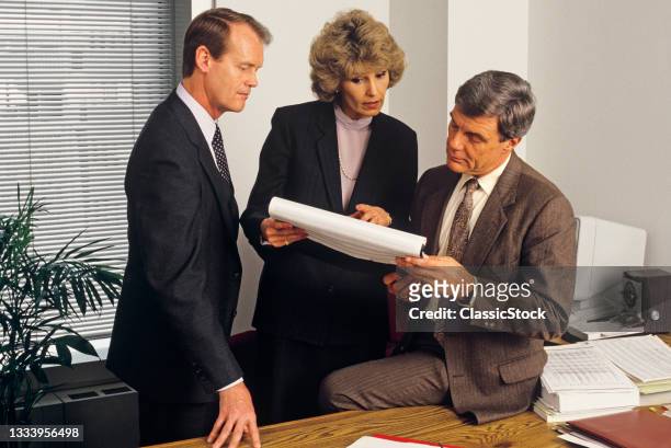 1980s Three Executives, Woman And Two Men, Holding Meeting In Office Reviewing Paper Work Spread Sheet.