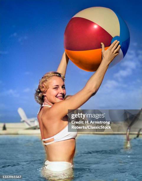 1950s Side View Of Blonde In White Bikini Standing In Pool In Waist-High Water Holding Beach Ball In Air .