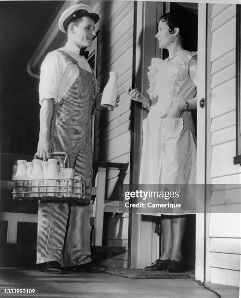 Milkman in uniform and hat with hand basket of dairy products making a residential delivery. Circa 1950.