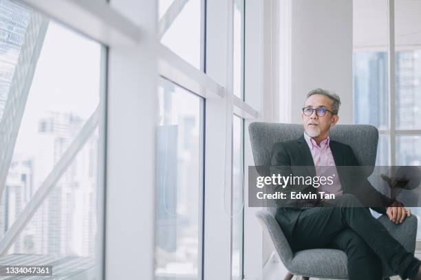 erfolgreicher asiatischer chinesischer geschäftsmann im anzug sitzt gekreuzt auf dem sofa in seinem bürozimmer mit blick auf die hauptstadt - asian man thinking stock-fotos und bilder