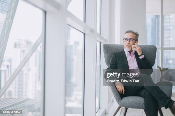 successful asian chinese senior businessman in suit sitting crossed leg on sofa in his office room listening smart phone - empire style furniture stock pictures, royalty-free photos & images