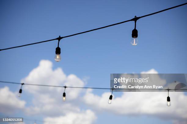 string lights against a blue sky with white puffy clouds - lichterkette sommer stock-fotos und bilder