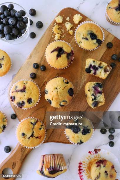 image of batch homemade blueberry muffins in paper cake cases on wooden chopping board, surrounded by fresh blueberries, marble effect background, elevated view - muffin stock pictures, royalty-free photos & images