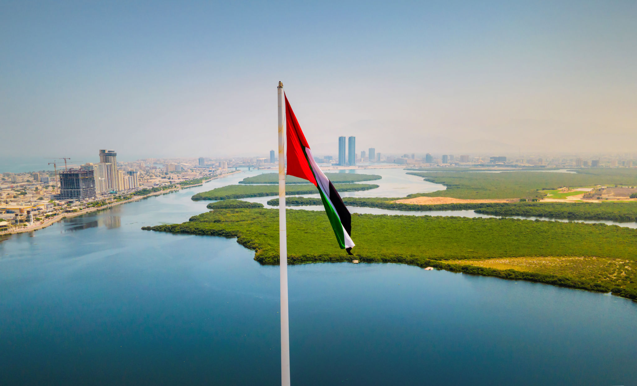 UAE national flag and Ras al Khaimah emirate aerial cityscape landmark skyline rising over the mangroves and the creek in the northern UAE UAE national flag and Ras al Khaimah emirate aerial cityscape landmark skyline rising over the mangroves and the creek in the northern UAE