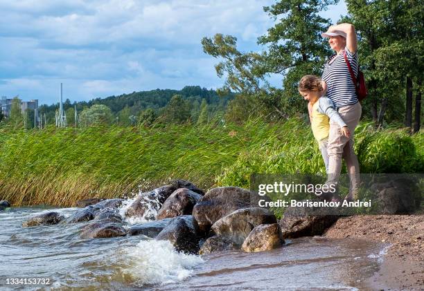 una joven y una niña caucásica de 6 años se paran en la orilla del lago y miran a lo lejos. un fuerte viento sopla en la cara y les encanta. - woman face beautiful 40 years fotografías e imágenes de stock