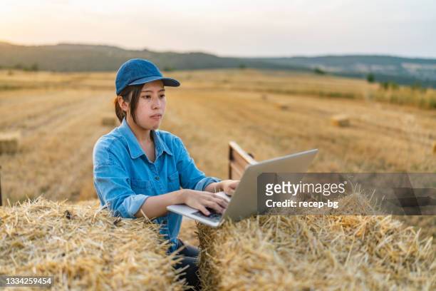 干し草の山のトラクタートレーラーに座って、農場でラップトップを使用して若い女性農家 - asian farmer ストックフォトと画像