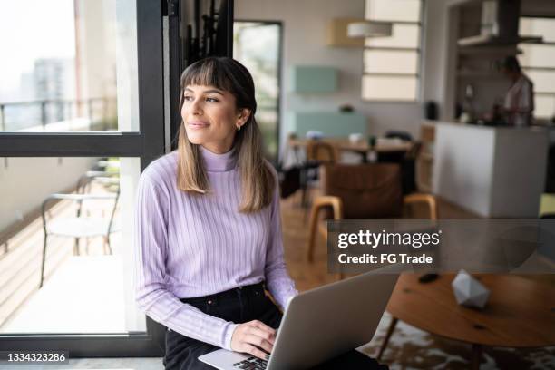 young woman sitting by the window sill looking away and using laptop at home - business hope stock pictures, royalty-free photos & images