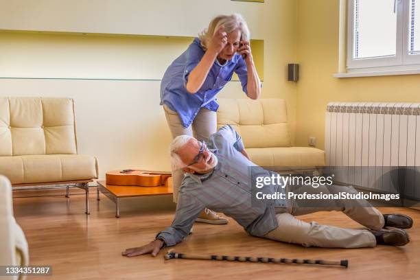 an older man with walking cane is lying on the floor after he had an accident, while his wife is helping him to get up and calling an urgent center. - patient lying down stock pictures, royalty-free photos & images