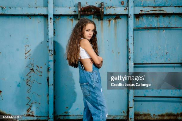 caucasian girl, dressed in a denim dungarees, posing, looking at the camera. with a blue metal street wall background. children, fashion and denim clothing concept. - preteen girl models stock pictures, royalty-free photos & images