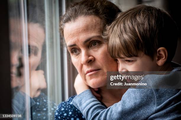 pensive mature woman posing with her son, very concerned looking through window worried about loss of her job and eviction due covid-19 pandemic - woman home with sick children stock pictures, royalty-free photos & images