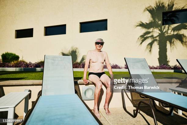 Wide shot portrait of senior male swimmer sitting on pool deck after early morning workout