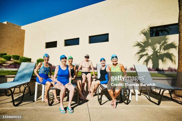 Wide shot portrait of group of smiling senior swimmers sitting on pool deck after morning workout