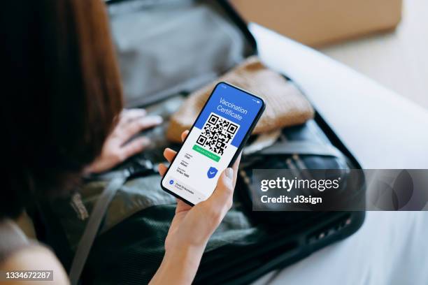 over the shoulder view of asian woman holding smartphone with a valid vaccination certificate for covid-19 for travel during the pandemic. packing her suitcase on bed preparing for travel in new normal - 疫苗護照 個照片及圖片檔