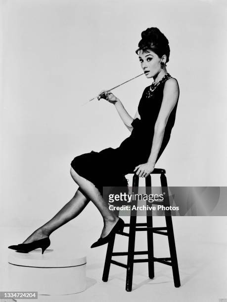 British actress Audrey Hepburn , wearing a black Hubert de Givenchy dress, holding a cigarette holder in character as Holly Golightly sitting on a...