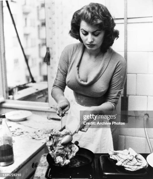Italian actress Sophia Loren, wearing a low-cut outfit and an apron, cooking in a kitchen, location unspecified, circa 1965.