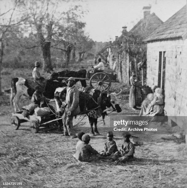 Group of young children sitting on the ground as women sit by a doorway and a man tends to a cattle-drawn cart in a scene of enslaved workers outside...