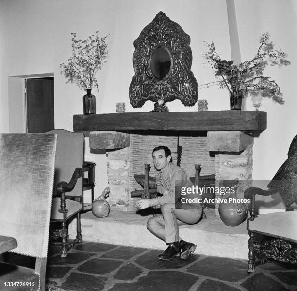 French-Armenian singer and actor Charles Aznavour sitting on the raised hearth of a fireplace, a large decorative mirror on the wall above the...