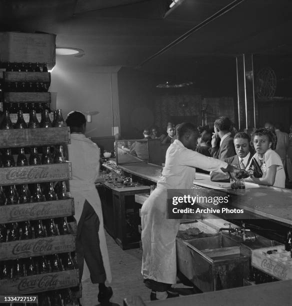 Crates containing bottles of Coca-Cola are stacked up behind the bar as white-coated bartenders serve drinkers at an unspecified bar, location...