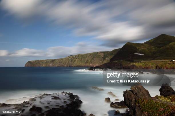 long exposure of calm sea seascape, rocks and cliffs under blue sky with moving clouds - fogo stockfoto's en -beelden