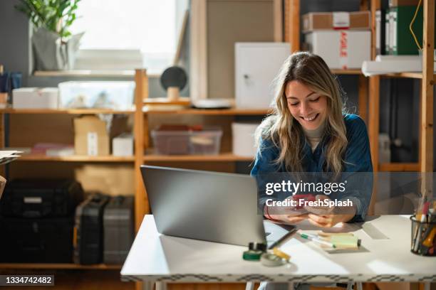 boosting productivity: young happy caucasian female entrepreneur sitting at her storage facility, multitasking on her computer and smartphone ( horizontal) - small business laptop bildbanksfoton och bilder