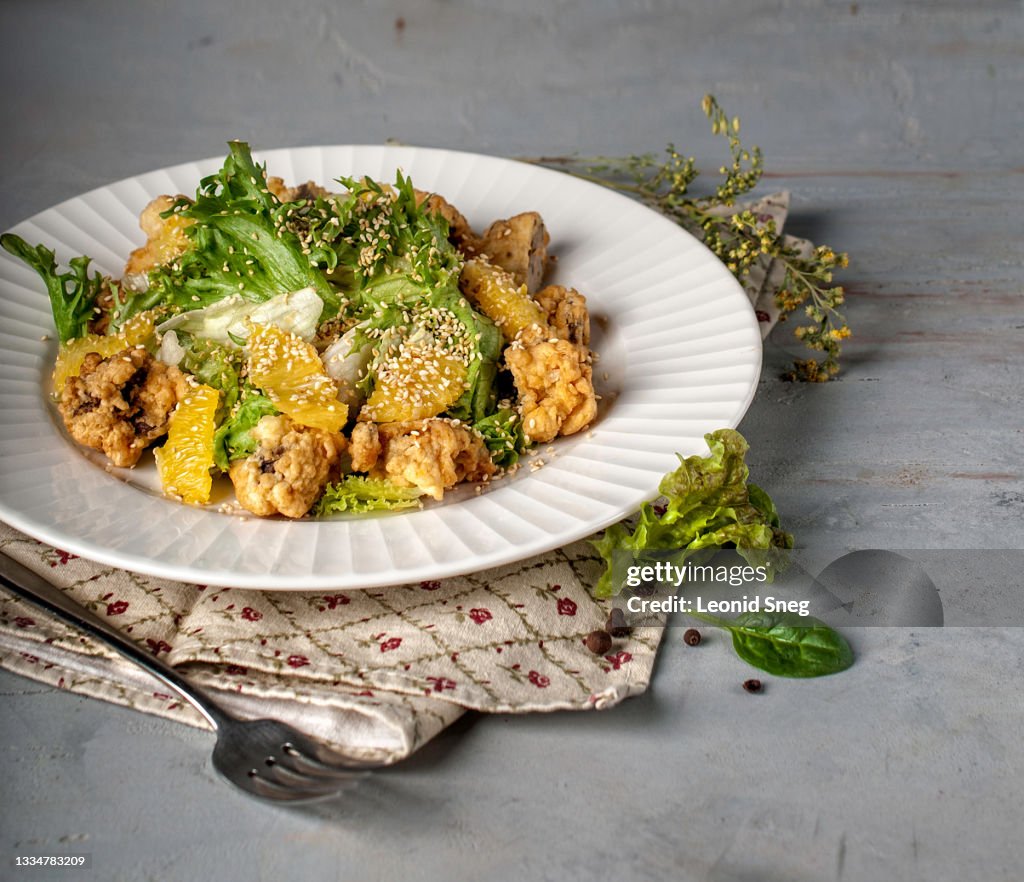 Food photography of green salad side view with deep fried chicken liver, fresh oranges and sesame seeds on a gray textured background close up