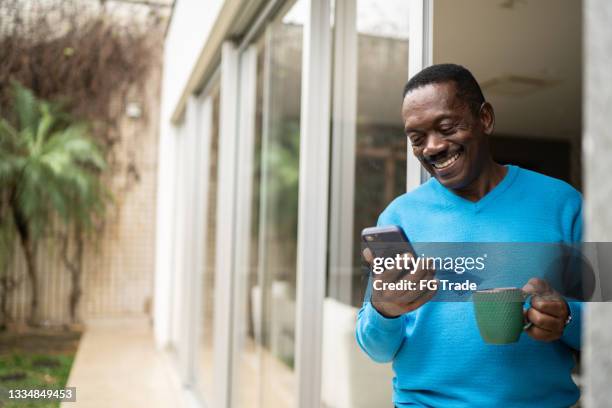 senior man holding coffee/tea mug and using smartphone at home - senior men coffee stock pictures, royalty-free photos & images