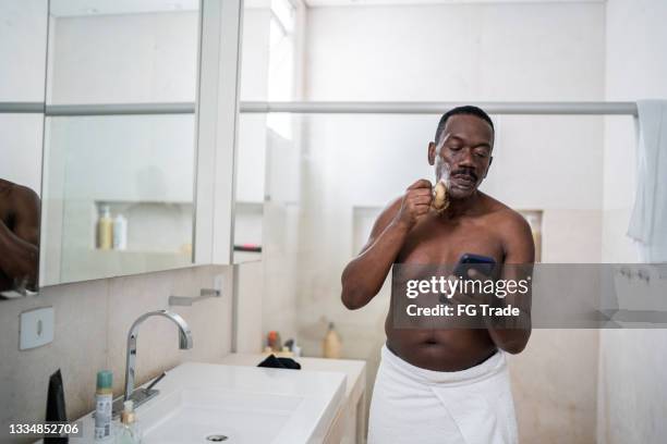 senior man applying shaving cream on face and using smartphone at home - man shaving foam stock pictures, royalty-free photos & images