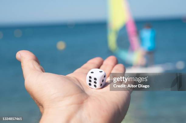 a person holds a dice at the edge of the beach. in the background and out of focus a person windsurfing. - chance photos et images de collection