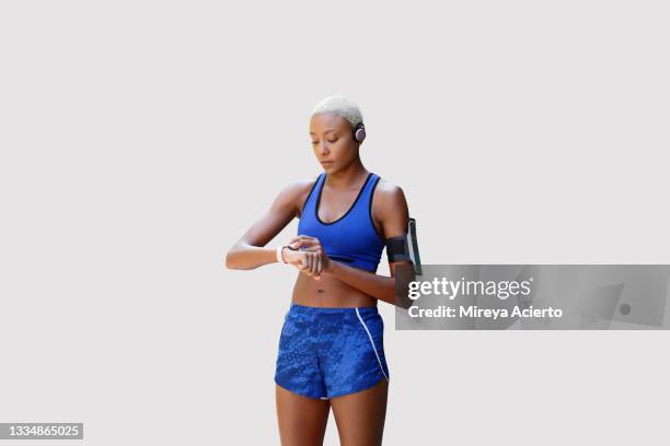 an african american millennial woman with short blond hair looks at her digital watch while wearing wireless headphones in front of a plain background wearing blue sports top and blue shorts. - running shorts stockfoto's en -beelden