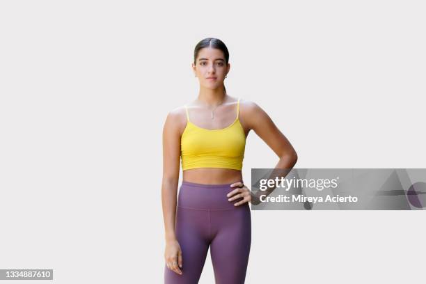 a latinx millennial woman with long hair stands confidently in front of a plain background wearing a yellow sports top and purple leggings. - leggings fotografías e imágenes de stock