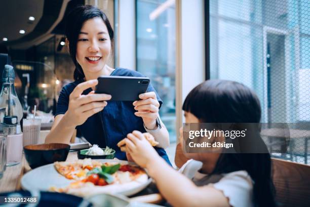young asian mother taking photos with smartphone of her lovely daughter while having lunch in restaurant. family enjoying a happy meal together. family lifestyle and technology - mother daughter brunch bildbanksfoton och bilder