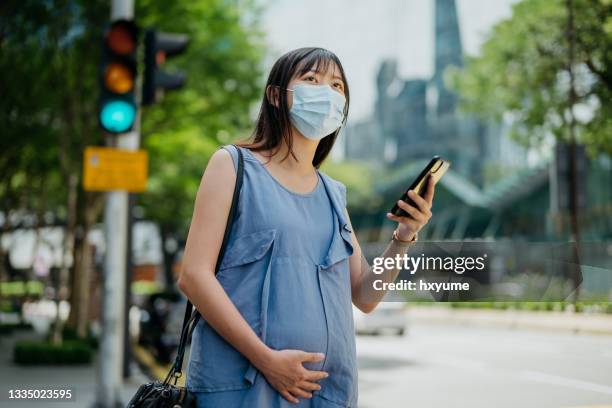 young pregnant woman with face mask hailing taxi at city street - pregnant woman car stock pictures, royalty-free photos & images