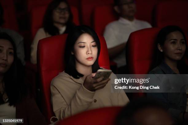asian chinese young woman using smart phone while movie is showing in movie theater. disturbing other audience around her - inconvenience stock pictures, royalty-free photos & images