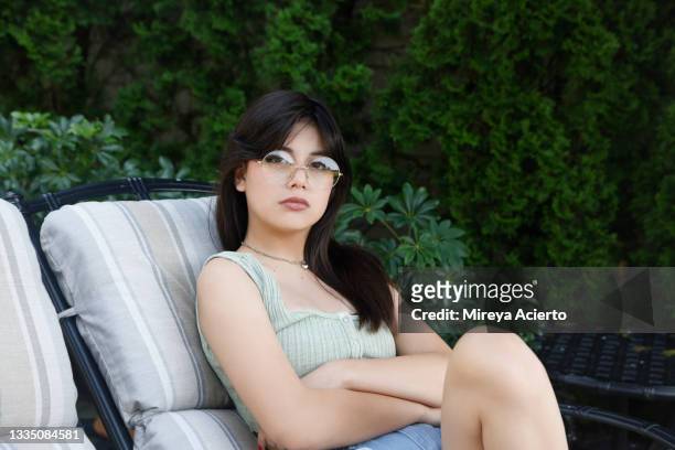 portrait of a mixed race generation z girl, with long hair, sits outside in the backyard, wearing round spectacles, a light green tank top and jean shorts. - 13 year old girls in shorts stock pictures, royalty-free photos & images