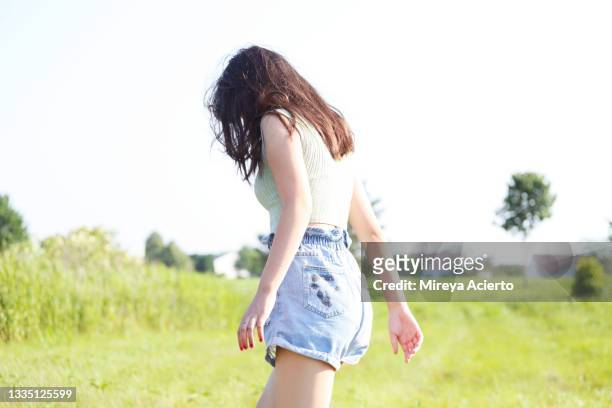 portrait of a mixed race generation z girl  with long hair, stretches her arms out in grassy field wearing a light green tank top and jean shorts. - 13 year old girls in shorts stock pictures, royalty-free photos & images
