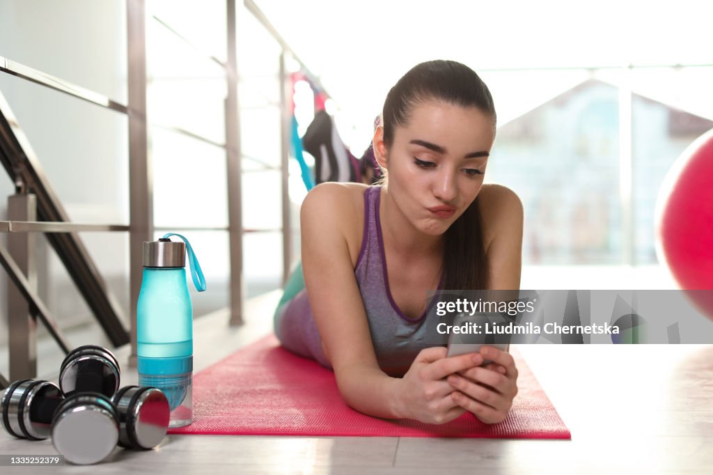 Lazy young woman with smartphone on yoga mat indoors