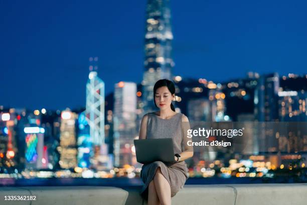 confident young asian businesswoman sitting on bench by the promenade of victoria harbour, using laptop on lap, against illuminated cityscape of hong kong at twilight in background - china banking regulatory commission stockfoto's en -beelden