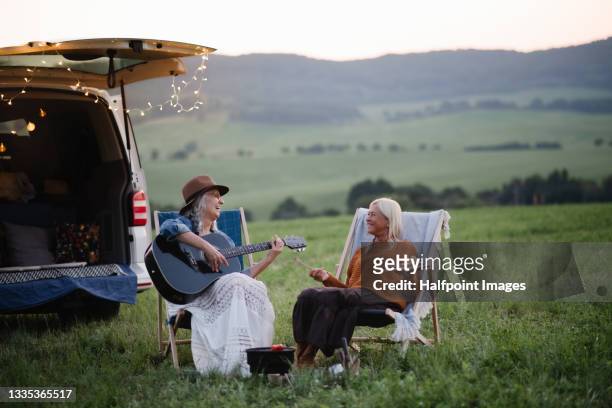 senior women friends sitting by car boot outdoors at sunset, caravan trip holiday. - woman playing guitar stock pictures, royalty-free photos & images