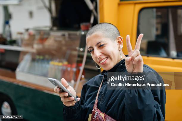 woman with shaved head giving peace symbol - woman entrepreneur looking at phone stock-fotos und bilder
