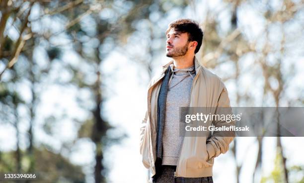 pensive young man standing alone in a park in the springtime - man jacket stock pictures, royalty-free photos & images