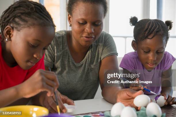 afro-american mother and daughters painting eggs for easter at living room table - pintura em têmpera imagens e fotografias de stock