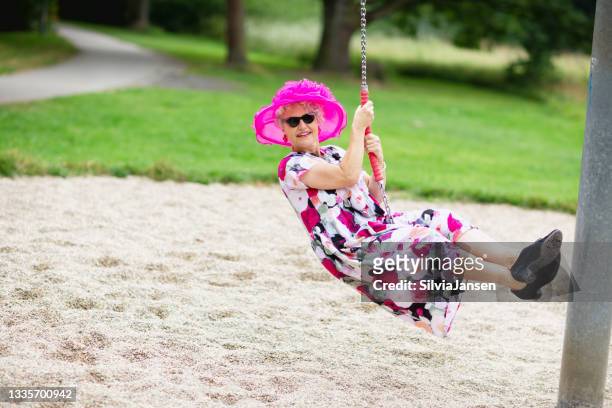 遊び場でスイングにピンクの帽子と花柄のドレスを着たエキセントリックなシニア女性 - eccentric ストックフォトと画像