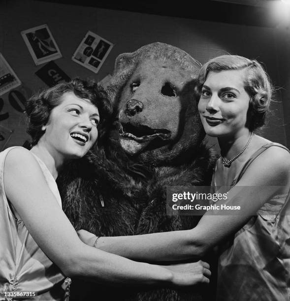 Two unspecified women embrace a person wearing a bear costume at a party, location unspecified, 1955.