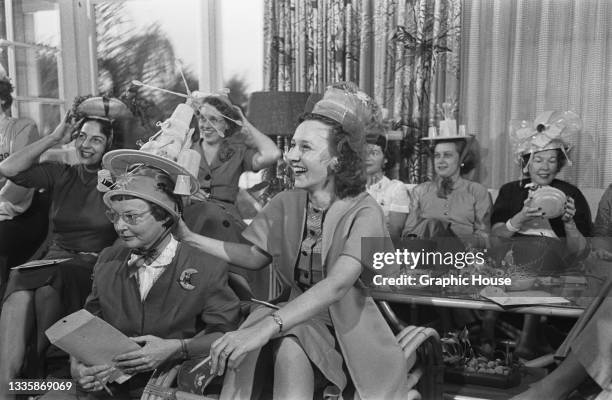 Group of unspecified women attend a Tupperware party, some wearing hats fashioned from Tupperware products, location unspecified, circa 1955....