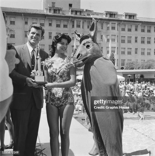 American politician Ted Kennedy presents a trophy to an unspecified woman wearing a swimsuit alongside a person dressed as a donkey, the mascot of...